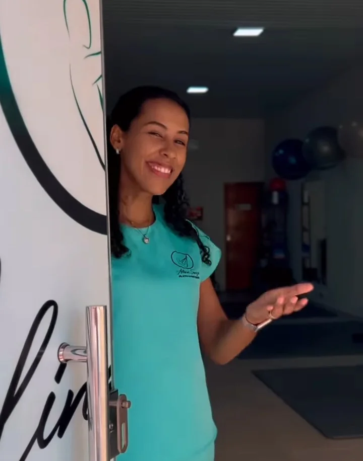 Aline Souza, de uniforme verde-água, abre a porta do Studio sorrindo e faz com a mão o gesto de quem convida a entrar